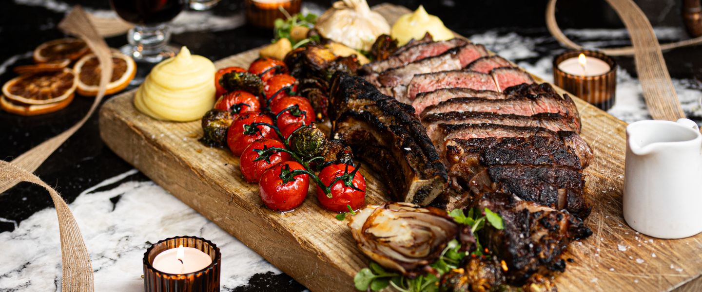 A wooden board with tomatoes on a vine, sliced steak, and mashed potatoes on it from Bread Street Kitchen