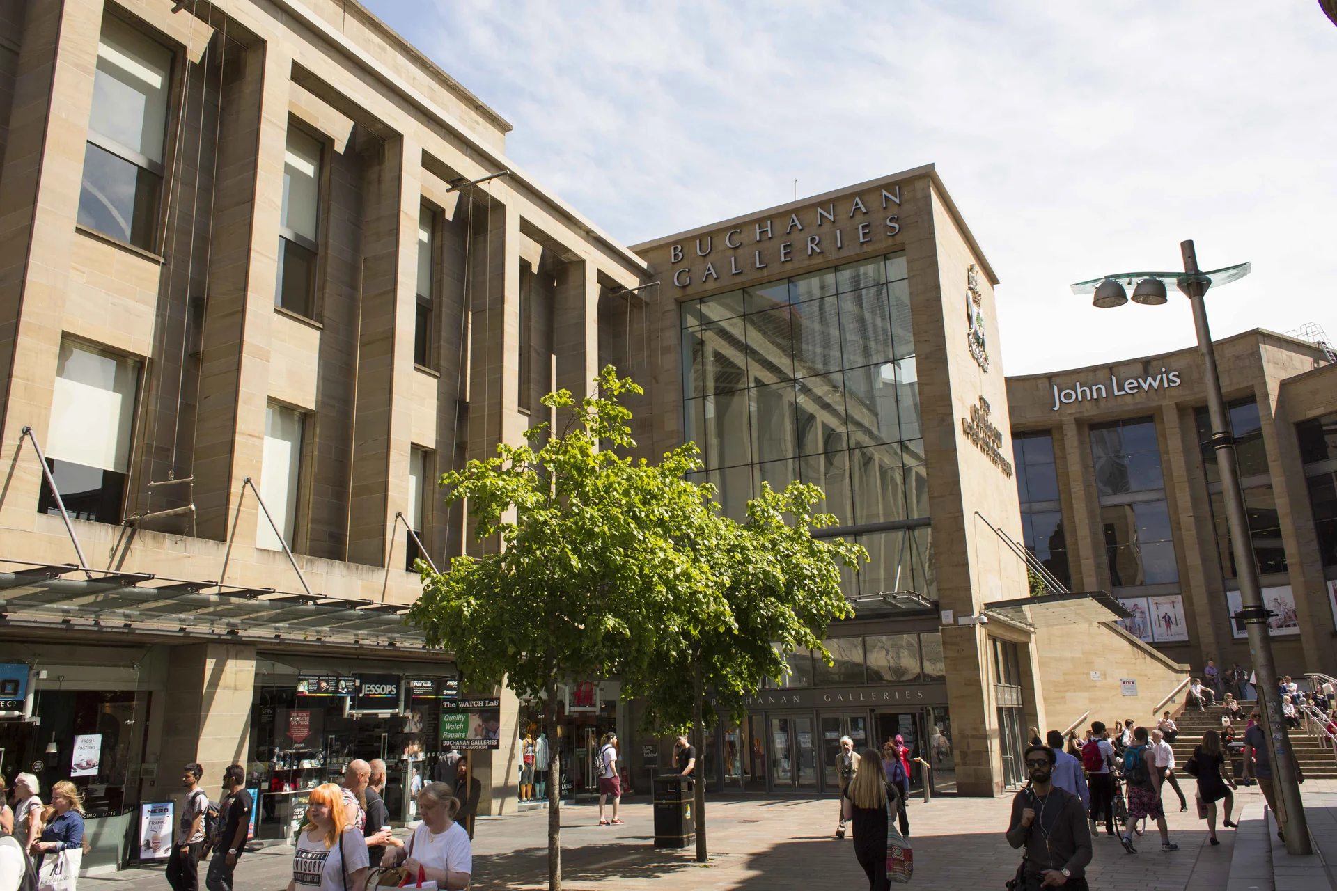 The main glass-front entrance of the **Buchanan Galleries** shopping centre in **Glasgow**, featuring the prominent signage and the adjacent **John Lewis** store, with shoppers navigating the busy pedestrianized plaza.