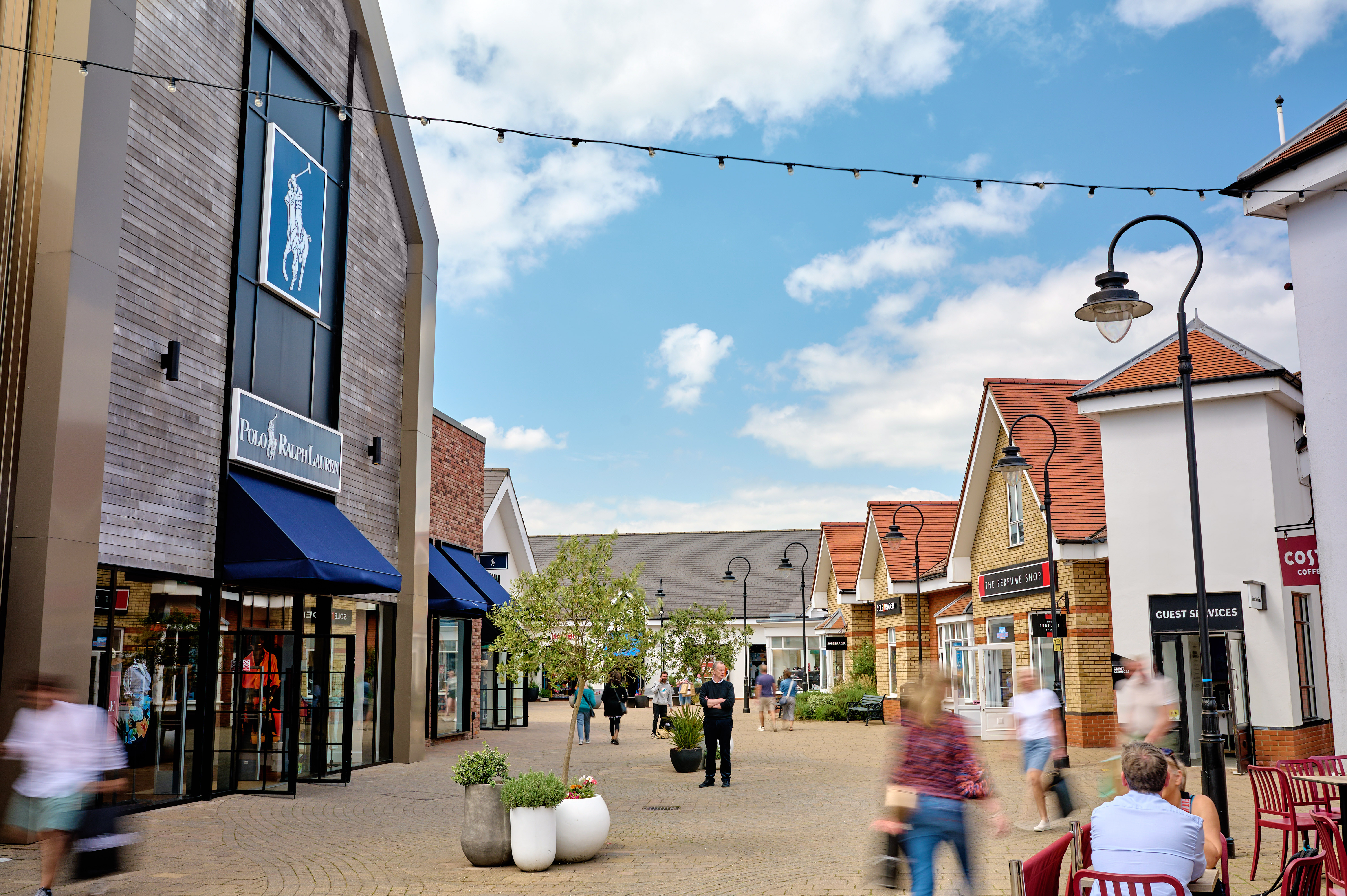 Braintree Village outlet shopping centre with an outdoor mall lined with various designer shops including Ralph Lauren. Guests are walking around carry shopping bags. There is a blue cloudy sky.