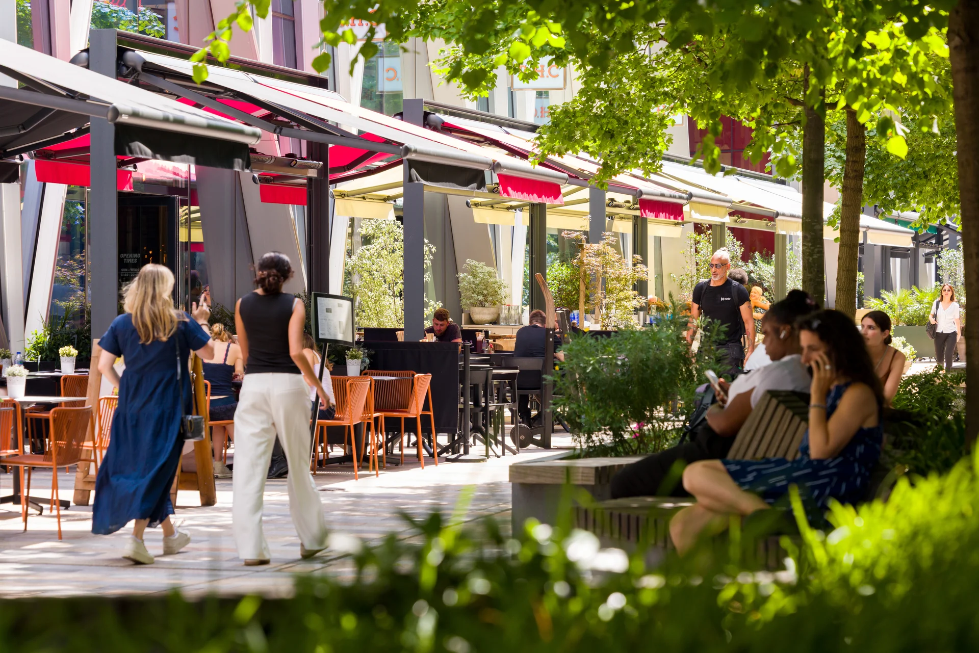 Outdoor café scene with people dining and walking, surrounded by greenery and modern buildings under a sunny sky.