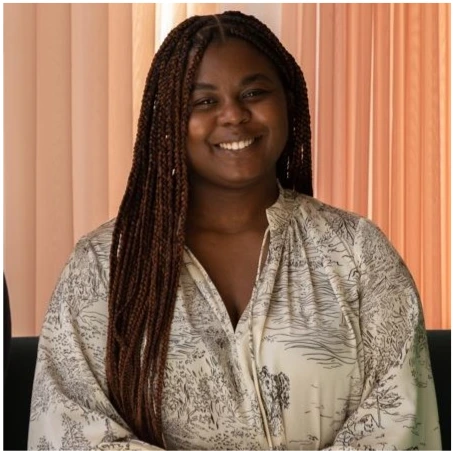 Person with braids in a patterned blouse sits before a pink curtain.