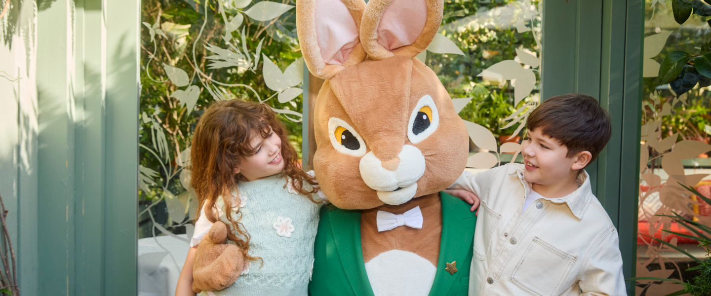 Two children smiling and posing with a large Easter bunny mascot wearing a green jacket and bow tie in a bright, plant-filled setting with decorative glass panels.