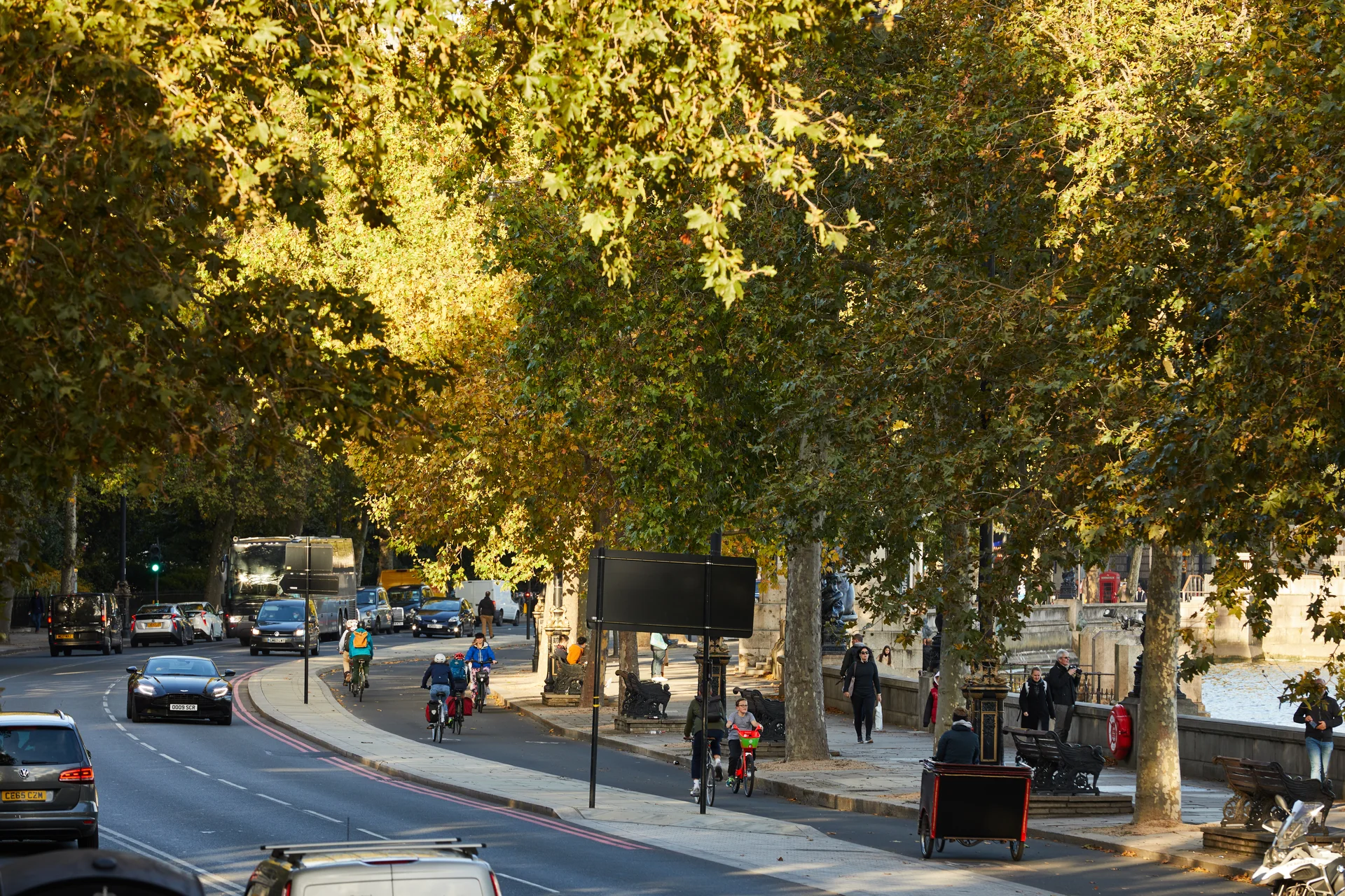 Street scene on the Strand in London, featuring heavy road traffic, cyclists in a dedicated lane, and pedestrians walking along the tree-lined embankment on a sunny autumn day.