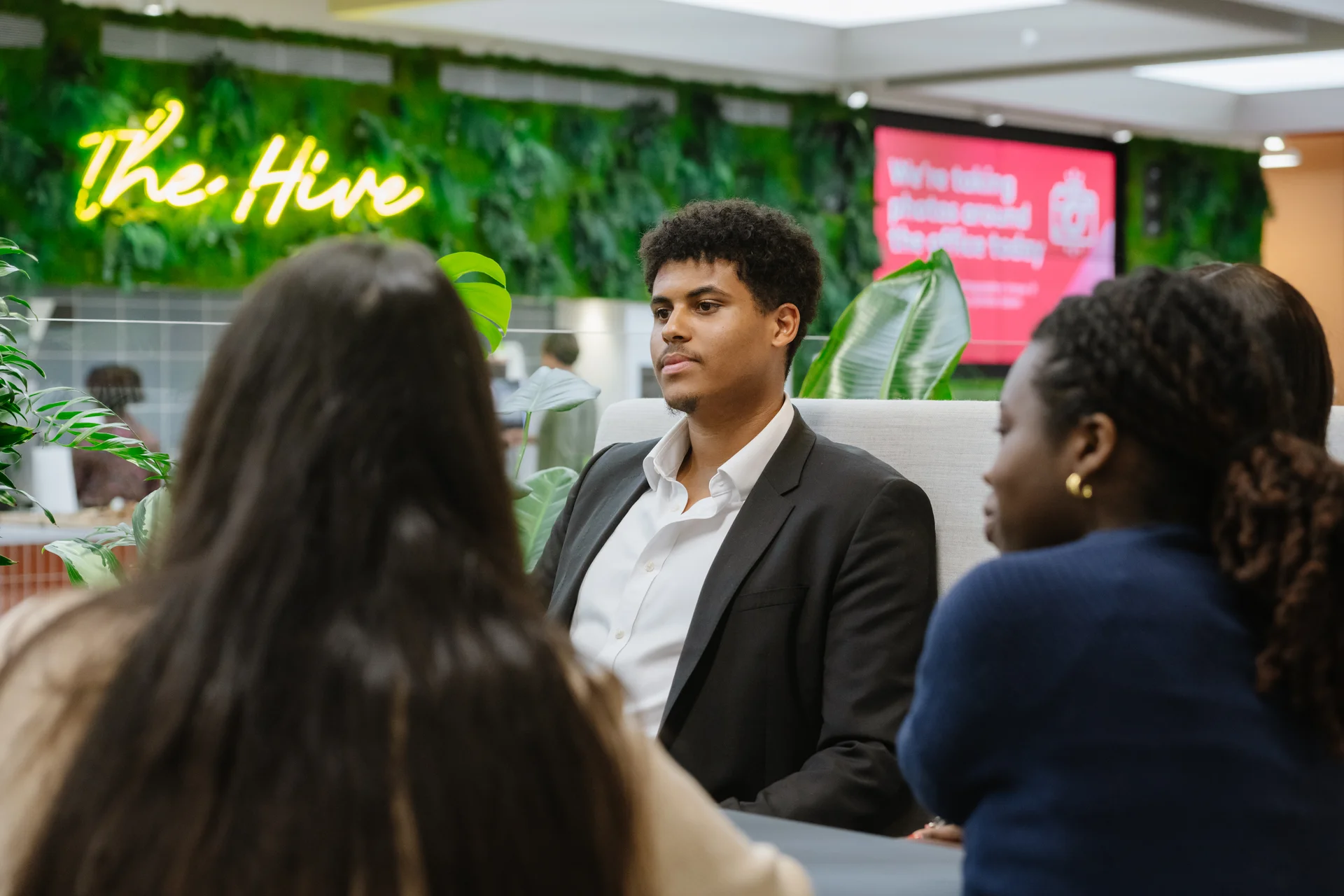 A group discusses in a modern office space with a green wall, neon 'The Hive' sign, and a digital display in the background.