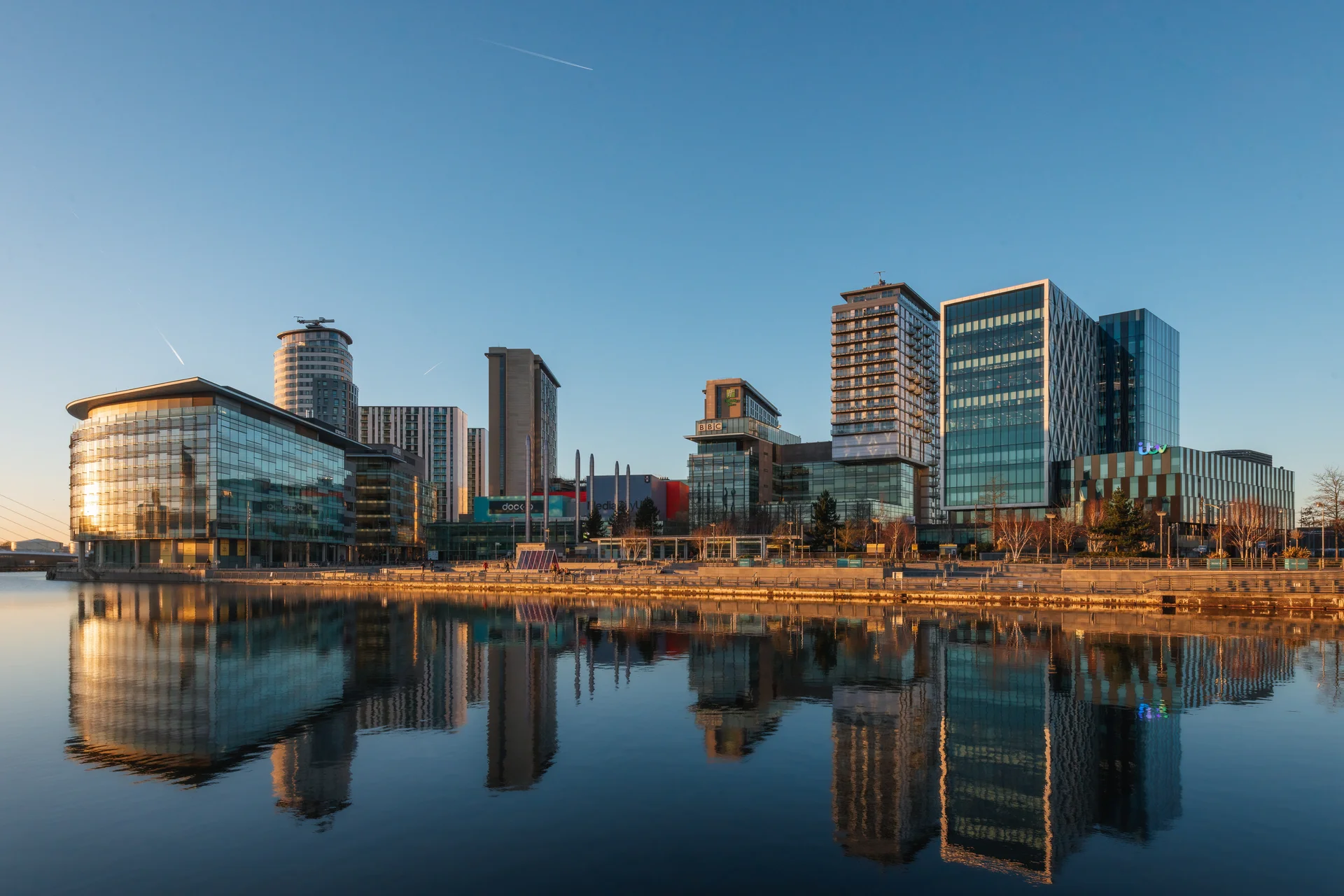 Modern glass buildings reflect in calm water under a clear blue sky, with a mix of architectural styles and heights.