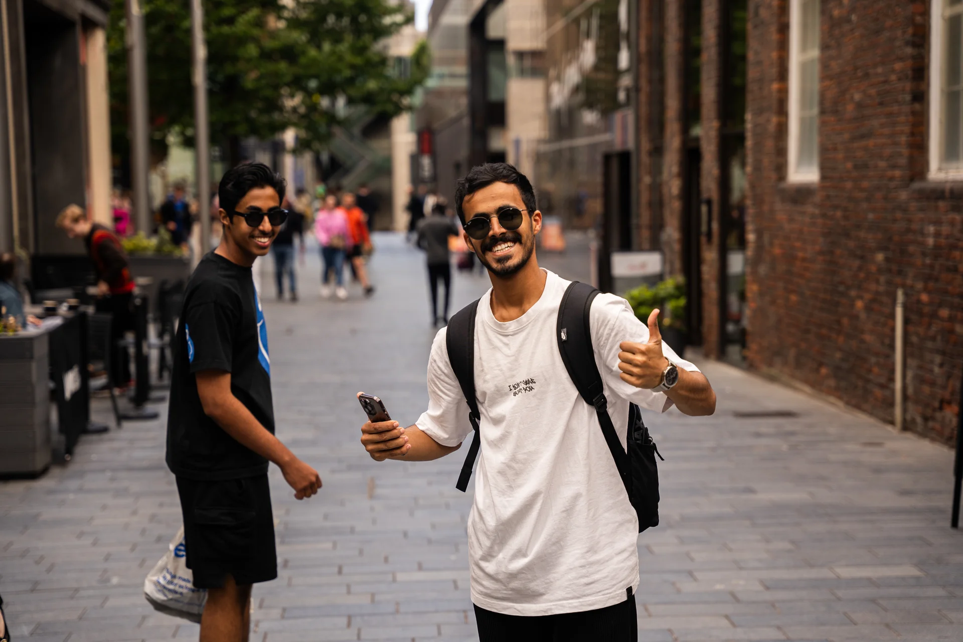 Two men walking on a paved street, one giving a thumbs-up, surrounded by brick buildings and greenery, daytime.