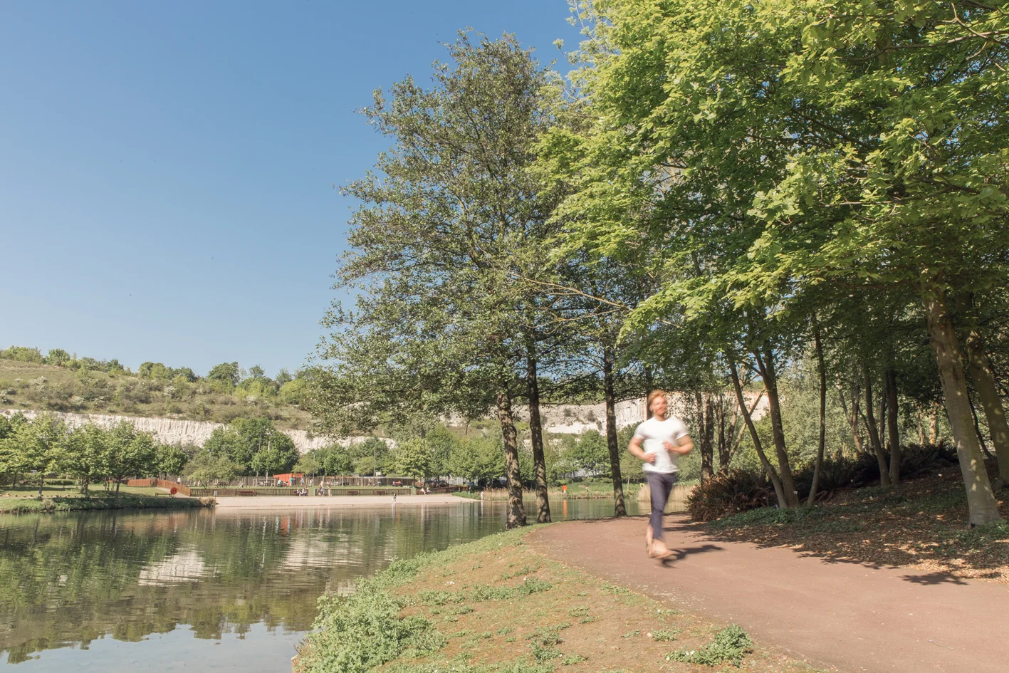 A person jogging on a path beside a calm lake, surrounded by lush green trees under a clear blue sky.