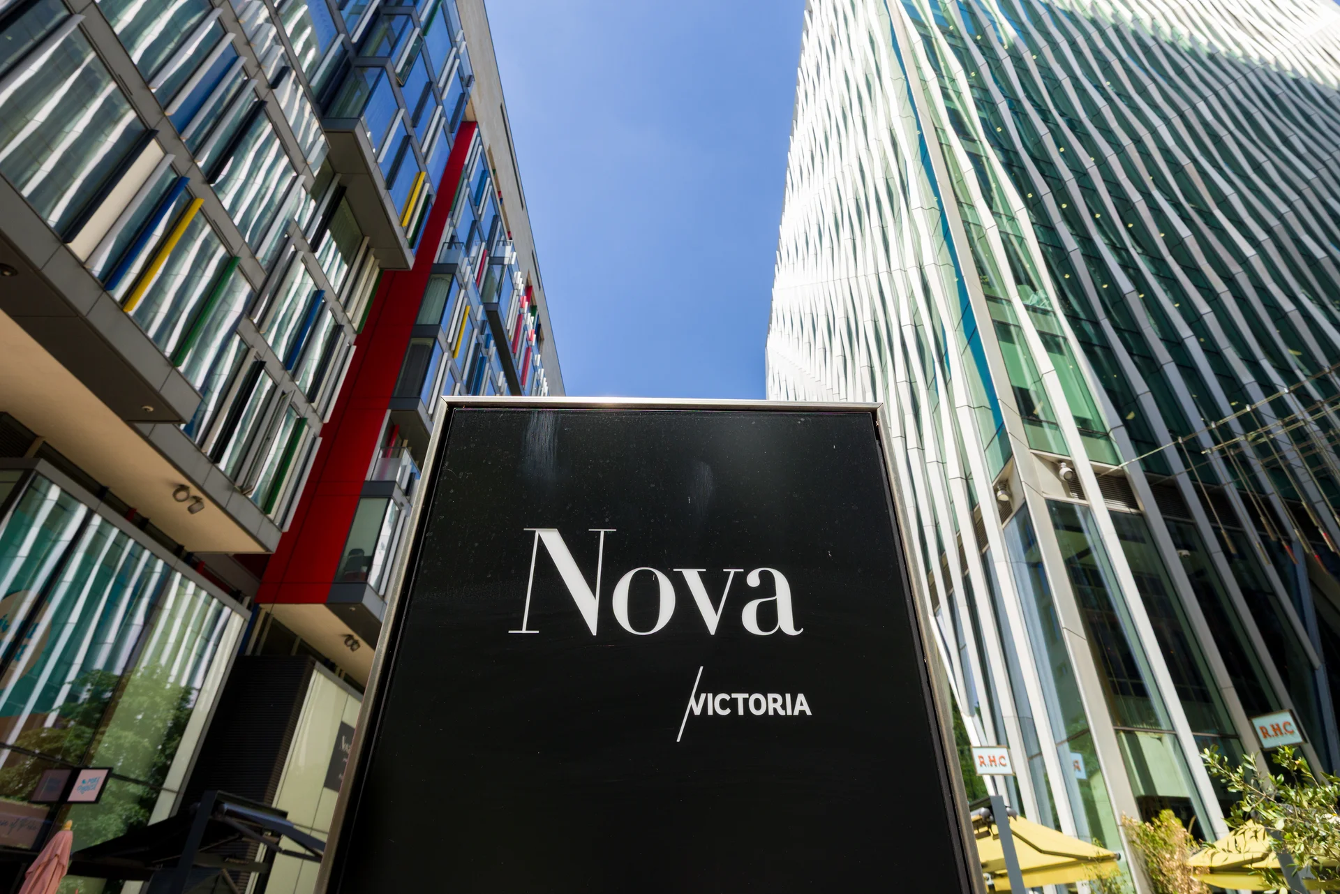 A modern urban scene with a 'Nova Victoria' sign, colorful glass buildings, and a clear blue sky.