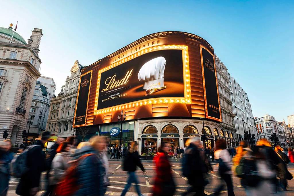 A bustling urban scene with a large, illuminated theater facade displaying a Lindt advertisement, surrounded by pedestrians.
