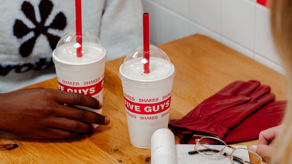 Two Five Guys milkshakes on a table next to some red leather gloves and a book
