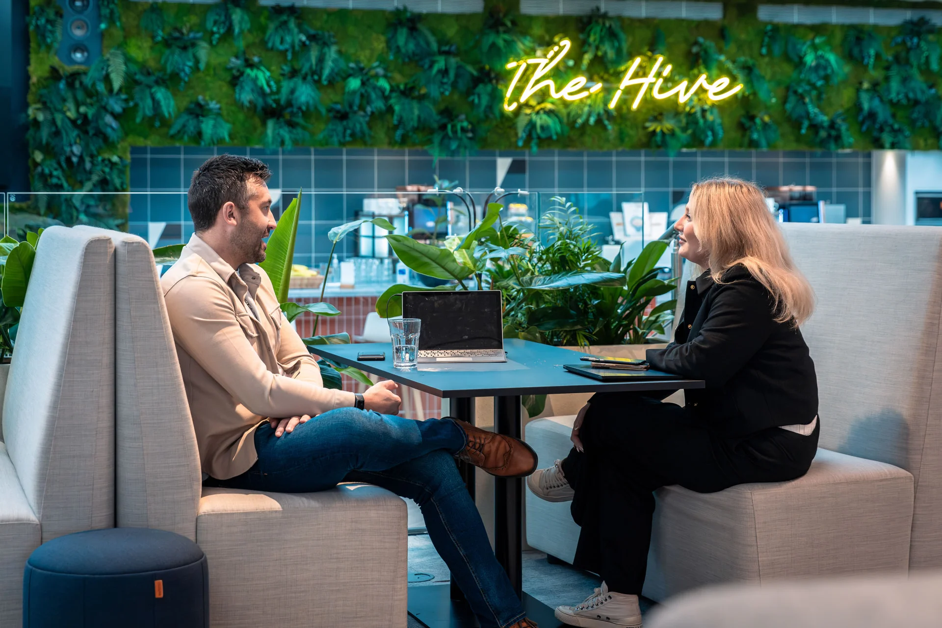 Two people converse at a modern café with greenery walls, neon 'The Hive' sign, and a cozy seating area.