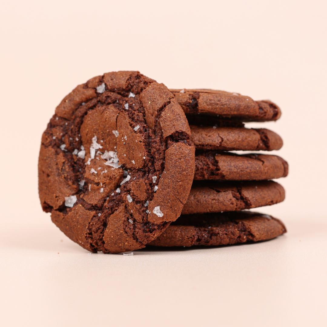 A stack of five brownie cookies with one extra cookie displayed face on leaning against the stack. chocolate colour sprinkled with sea salt on a pale pink background