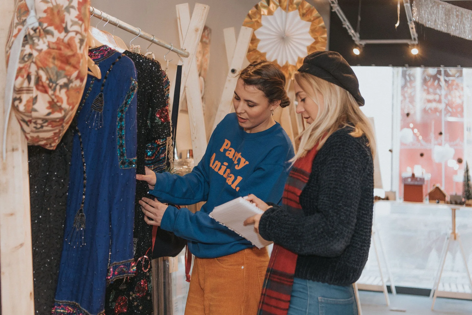 Two women browse clothing in a boutique with festive decorations, one holding a clipboard, under warm indoor lighting.