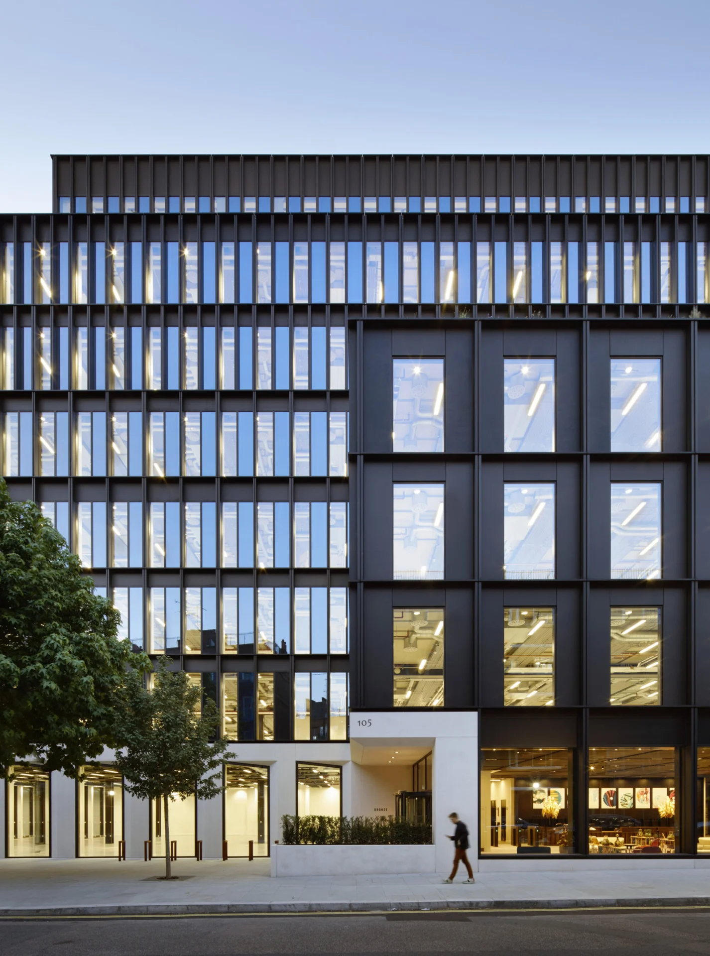 Modern multi-story building with black facade, large windows, and illuminated interior. A pedestrian walks on the sidewalk below.