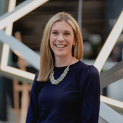 Jennie Colville in blue top and silver necklace standing before a geometric light structure.