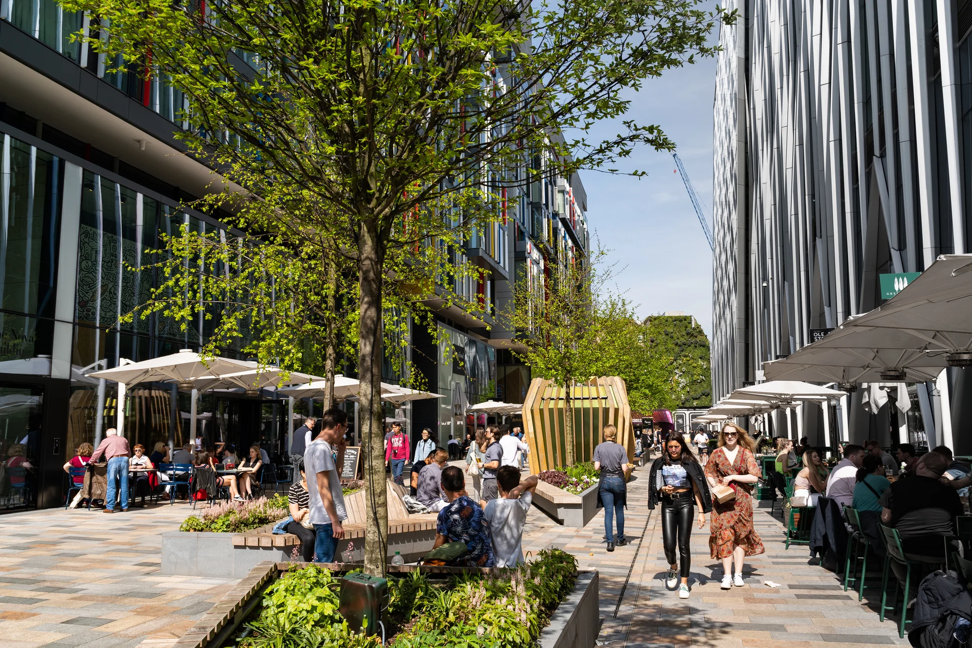 Outdoor plaza between glass buildings with trees, benches, and people dining