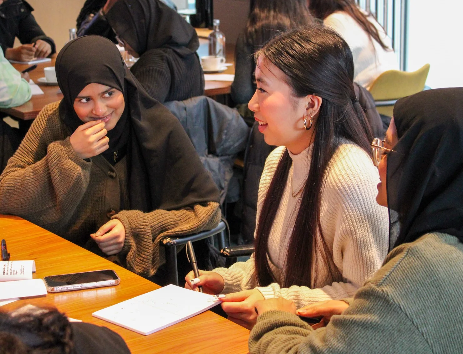 Three women engaged in conversation at a wooden table, with one writing notes and another smiling, in a casual indoor setting.