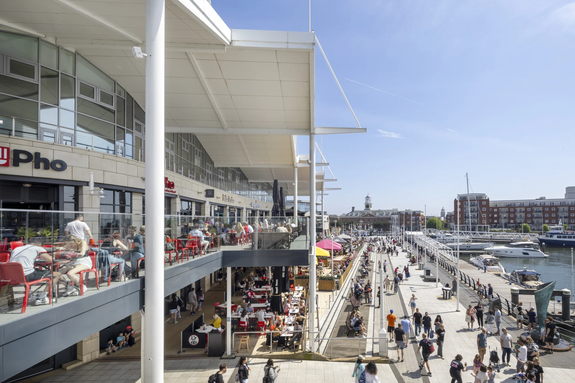 Elevated view of the waterfront at **Gunwharf Quays**, Portsmouth, showing the outdoor dining terraces of restaurants like Pho and a busy pedestrian promenade along the marina on a sunny day.