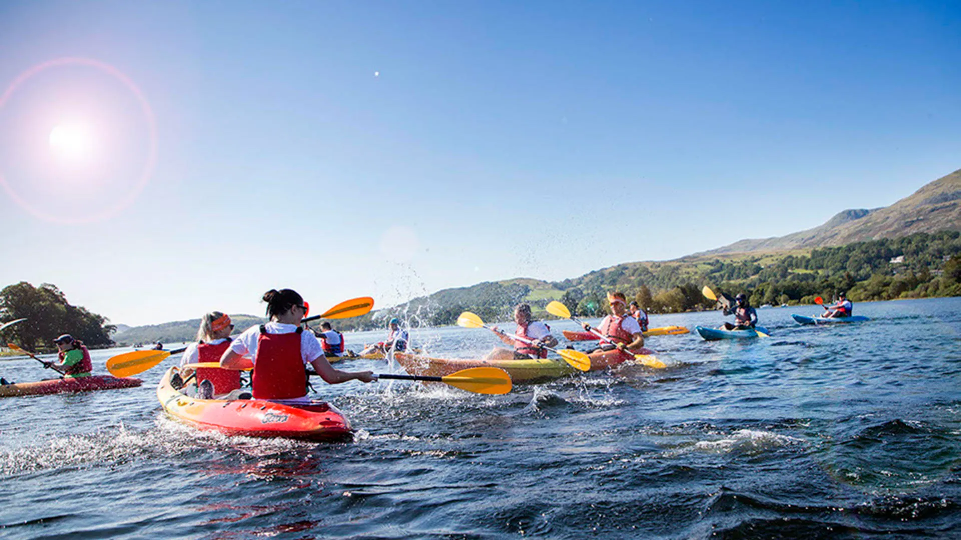 Group kayaking on a calm lake with green hills under a sunny blue sky.