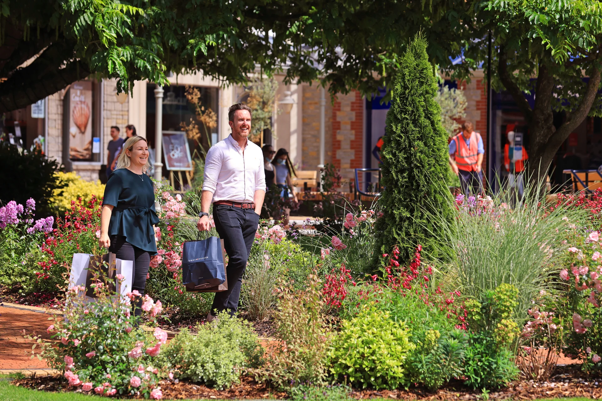 A smiling couple walking past large, vibrant flower beds and established shade trees at the outdoor **Clarks Village** outlet shopping centre.