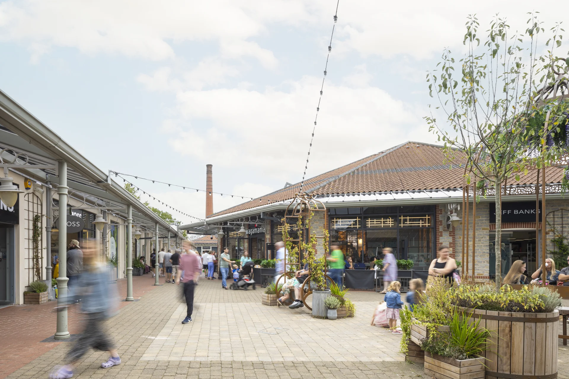 A sunny view of the dining area at **Clarks Village**, showing people seated at outdoor tables next to a building with a red tile roof and a tall brick chimney, with shoppers passing on the paved walkway.