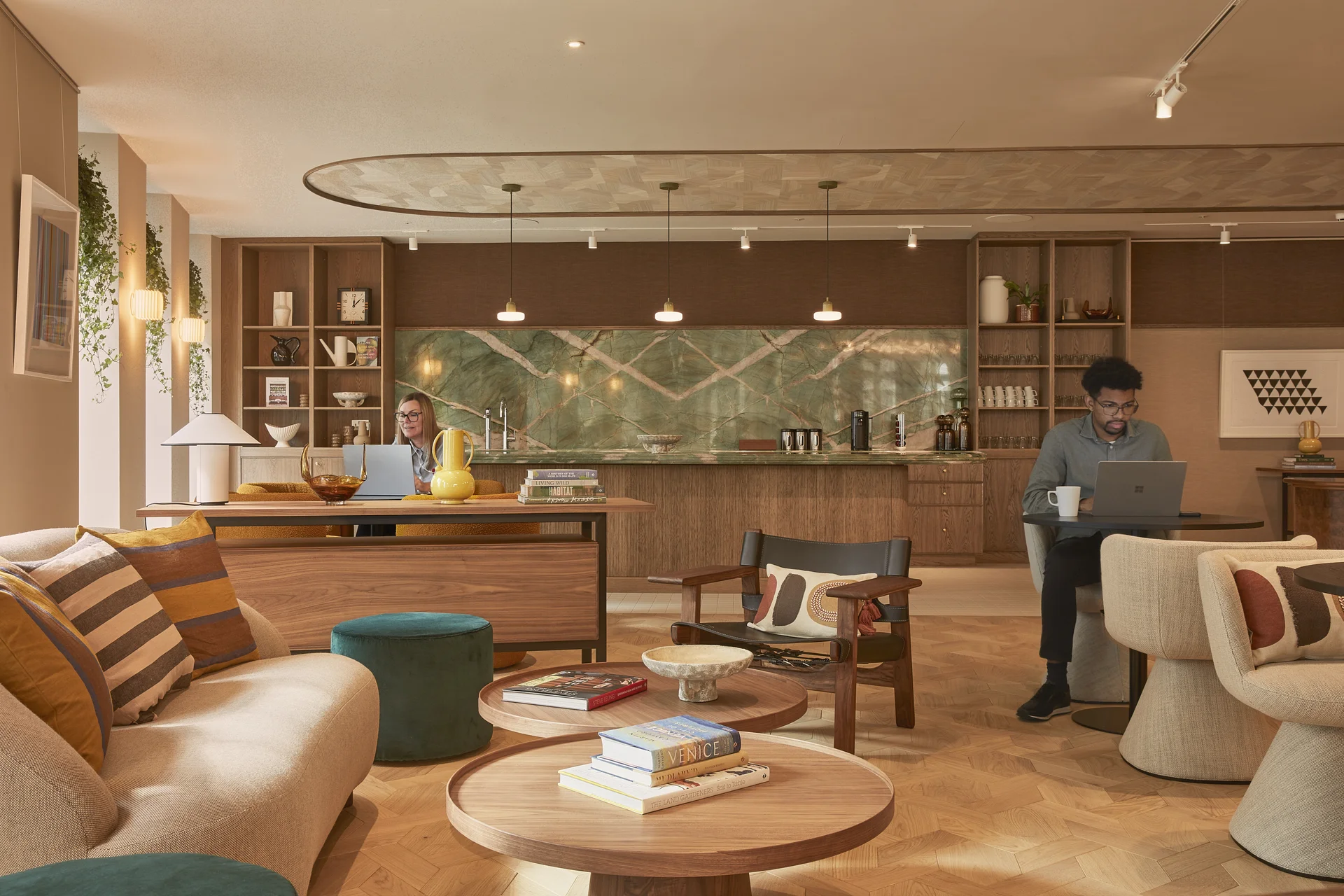 Modern lounge with wooden floors, cozy seating, a bar area with green marble backsplash, and people working on laptops.