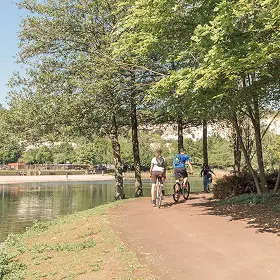 Two cyclists and a child riding on a dirt path next to a tree-lined lake on a sunny day, with a leisure area and cliff face visible in the background.