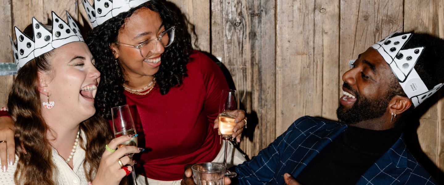 Three people holding wine glasses and wearing silver Christmas hats laughing around a table
