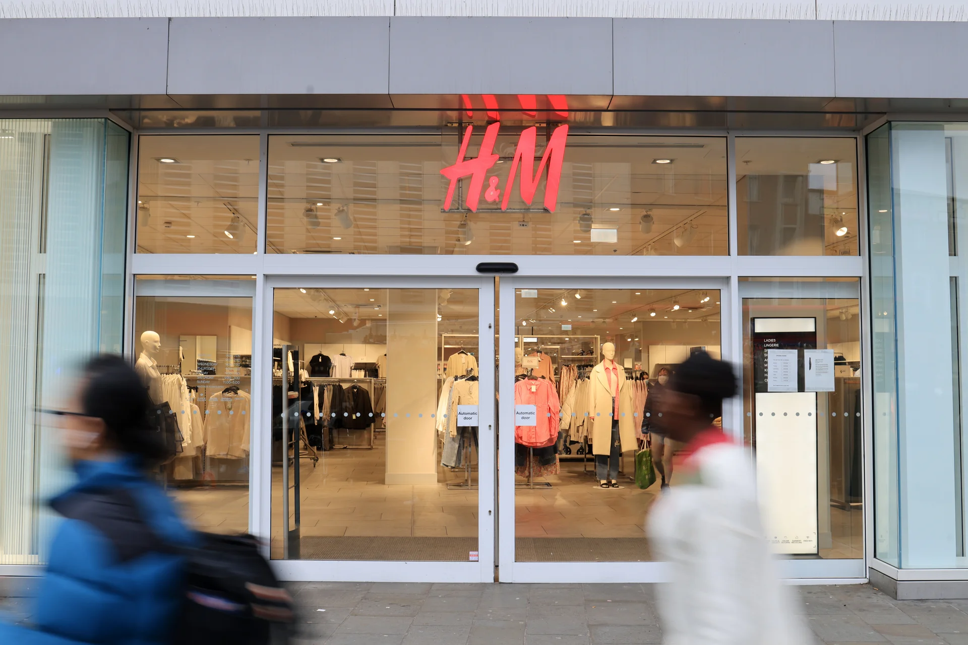 Close-up of the H&M storefront entrance at Lewisham Shopping Centre. Blurry shoppers walk past the large windows.