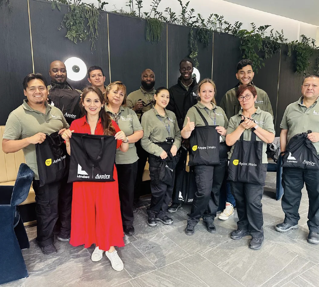 Diverse team of ten employees in green and black uniforms posing with a woman in red, holding black partnership tote bags.