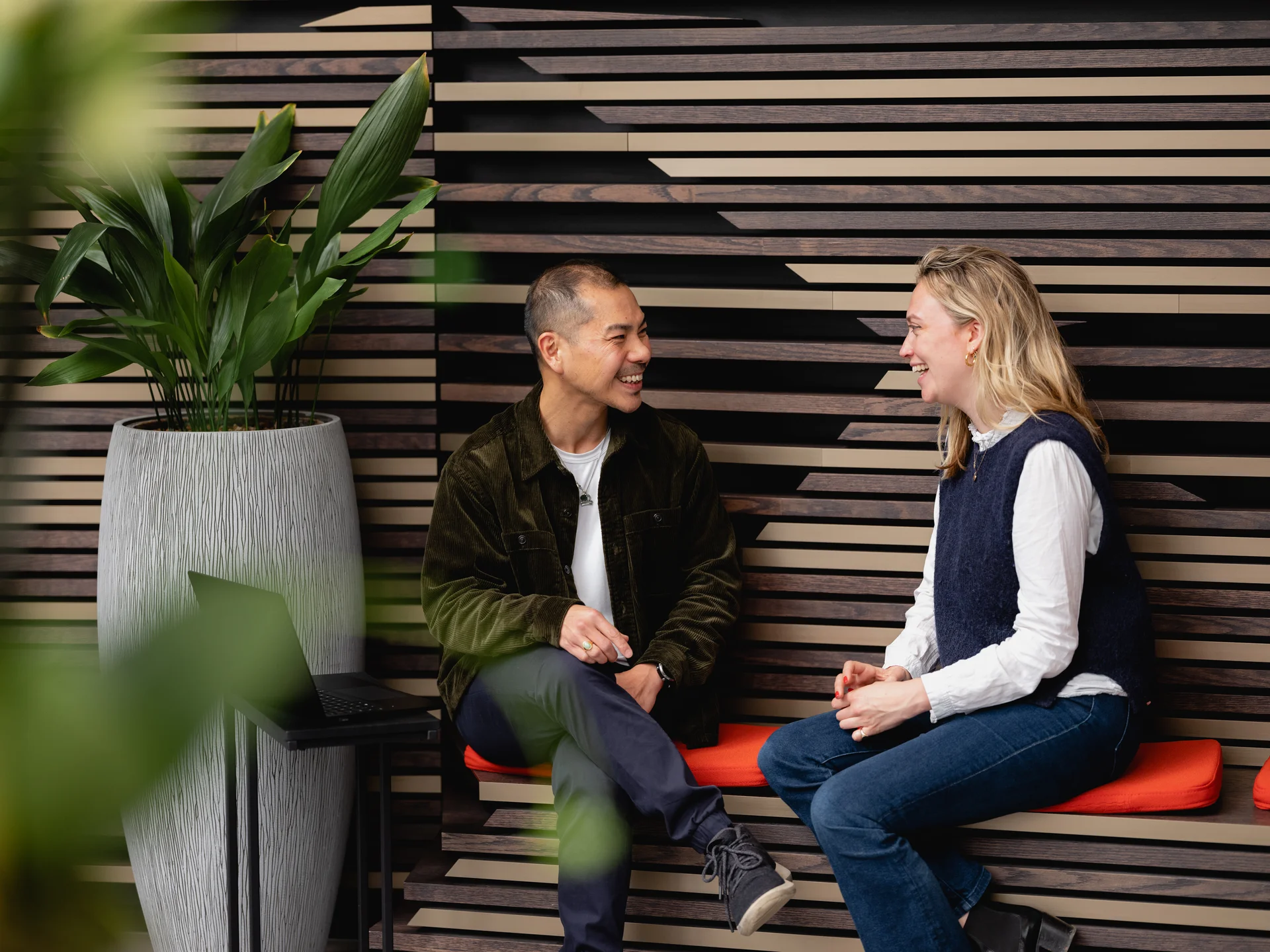 Two people sit on a wooden bench with a striped wall backdrop, conversing. A laptop and a potted plant are nearby.