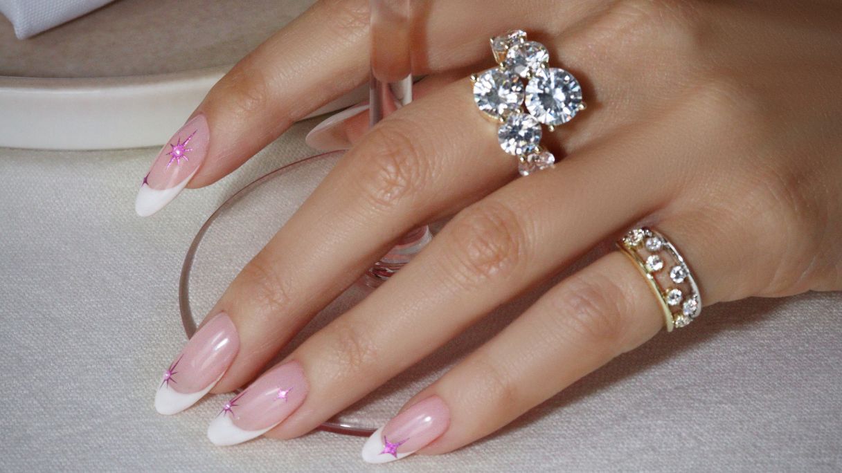 A hand with a French manicure with pink shimmery sparkles wearing chunky rings holding the base of a glass of wine