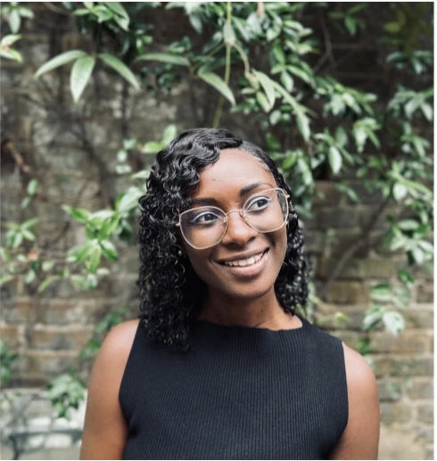 Person with curly hair and glasses smiling in a black top, with green foliage and brick wall background.