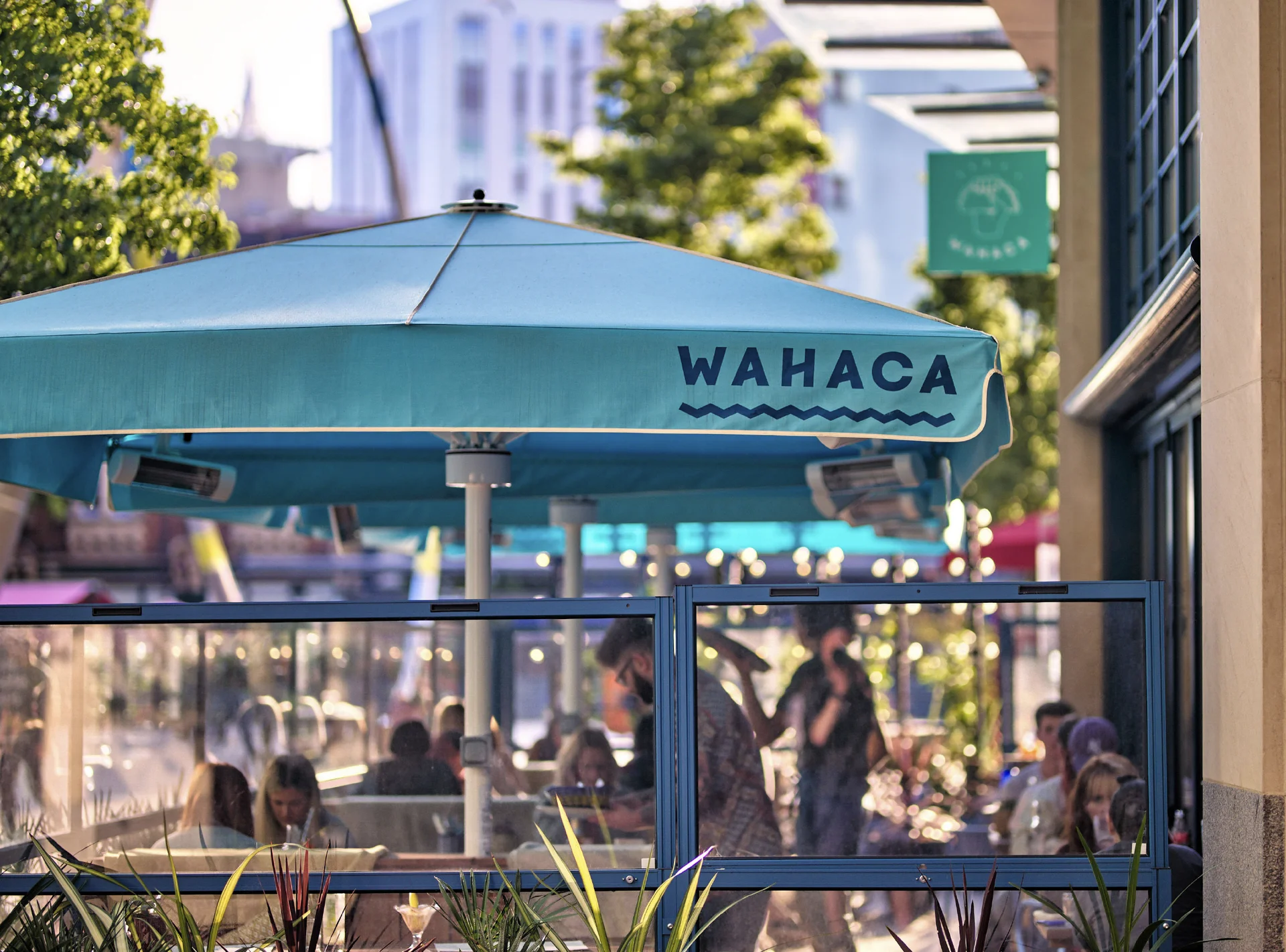 People eating at an outdoor restaurant terrace with a **Wahaca** umbrella on a sunny day with city buildings in the background.