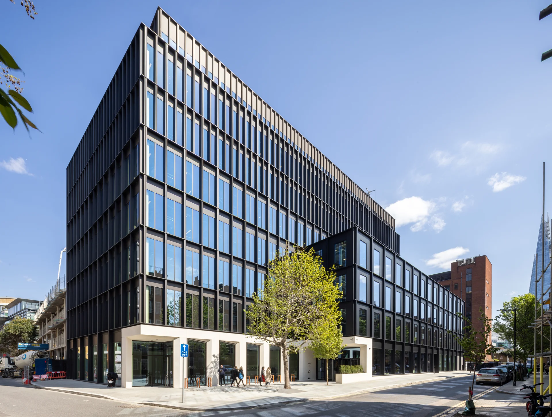 Modern multi-story building with black facade and glass windows, surrounded by trees and pedestrians on a sunny day.