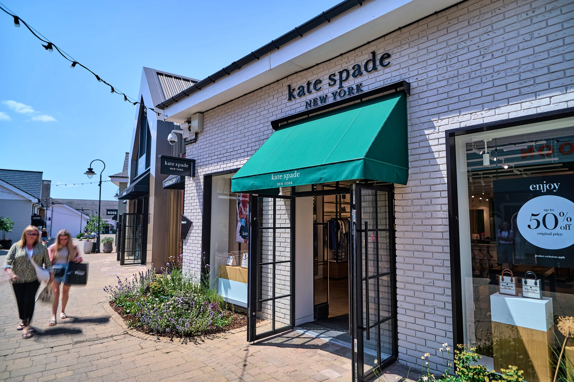 The **Kate Spade New York** storefront at **Braintree Village**, featuring a white brick facade and a bright green awning, with shoppers walking past outdoor planters on a bright day.