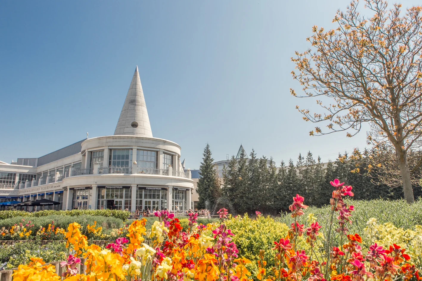 Exterior view of the Bluewater shopping center building with its iconic conical roof, framed by vibrant yellow, pink, and red spring flowers against a bright blue sky.