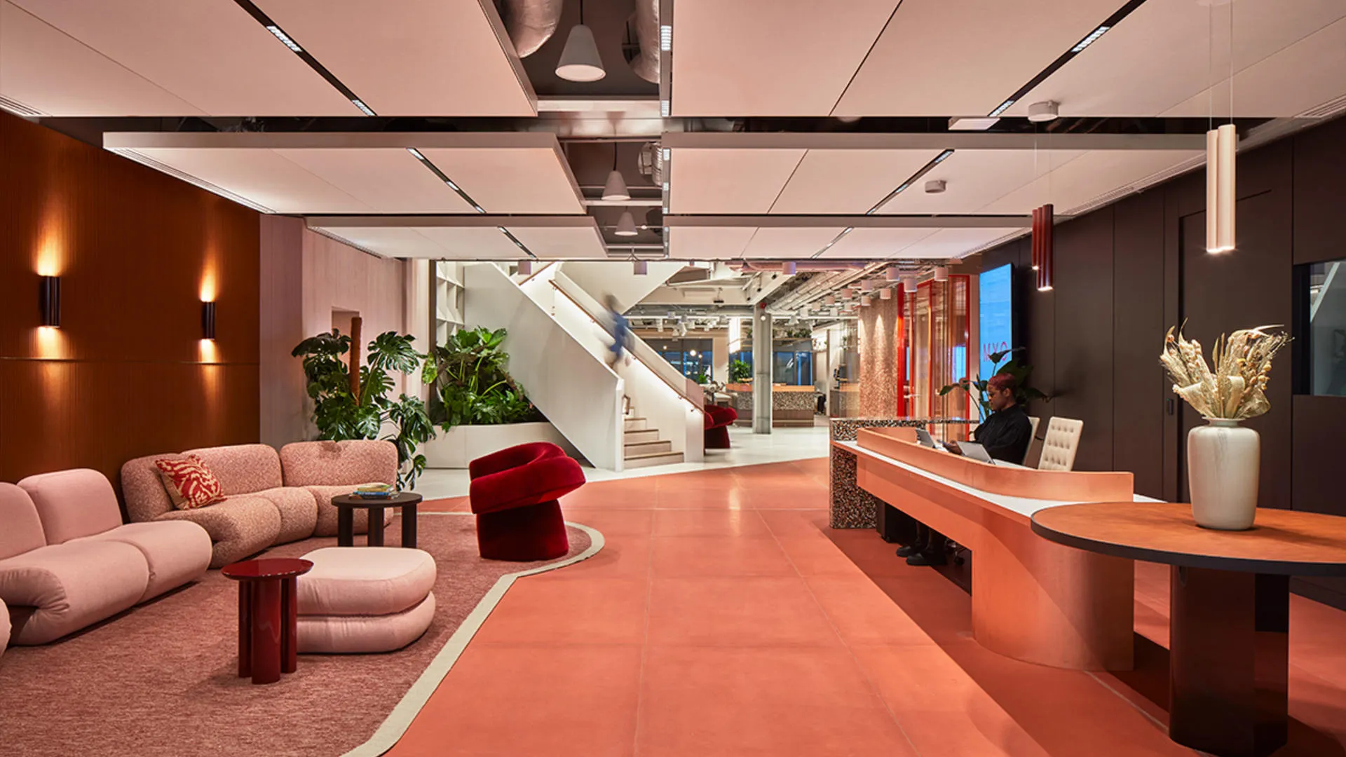 Modern office reception with pink and orange decor, sofa, armchair, plants, and a reception desk.