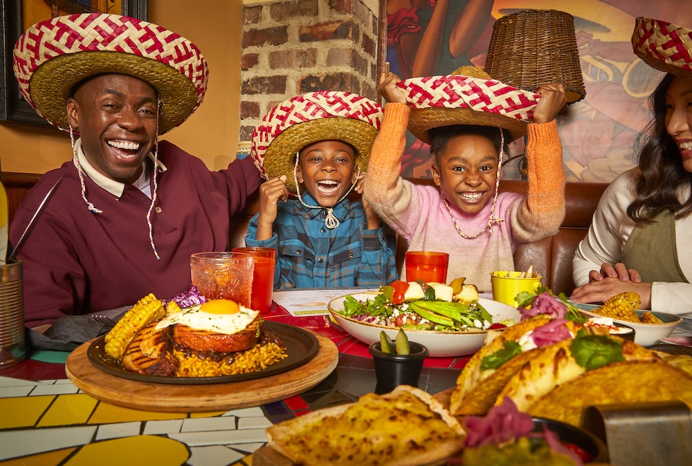 A family consisting of two adults and two children gathered a table with classic Mexican dishes wearing Sombrero hats and smiling