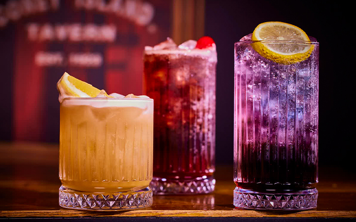 A selection of three cocktails, orange with a wedge of orange, red with a cherry and purplr with a slice of lemon 