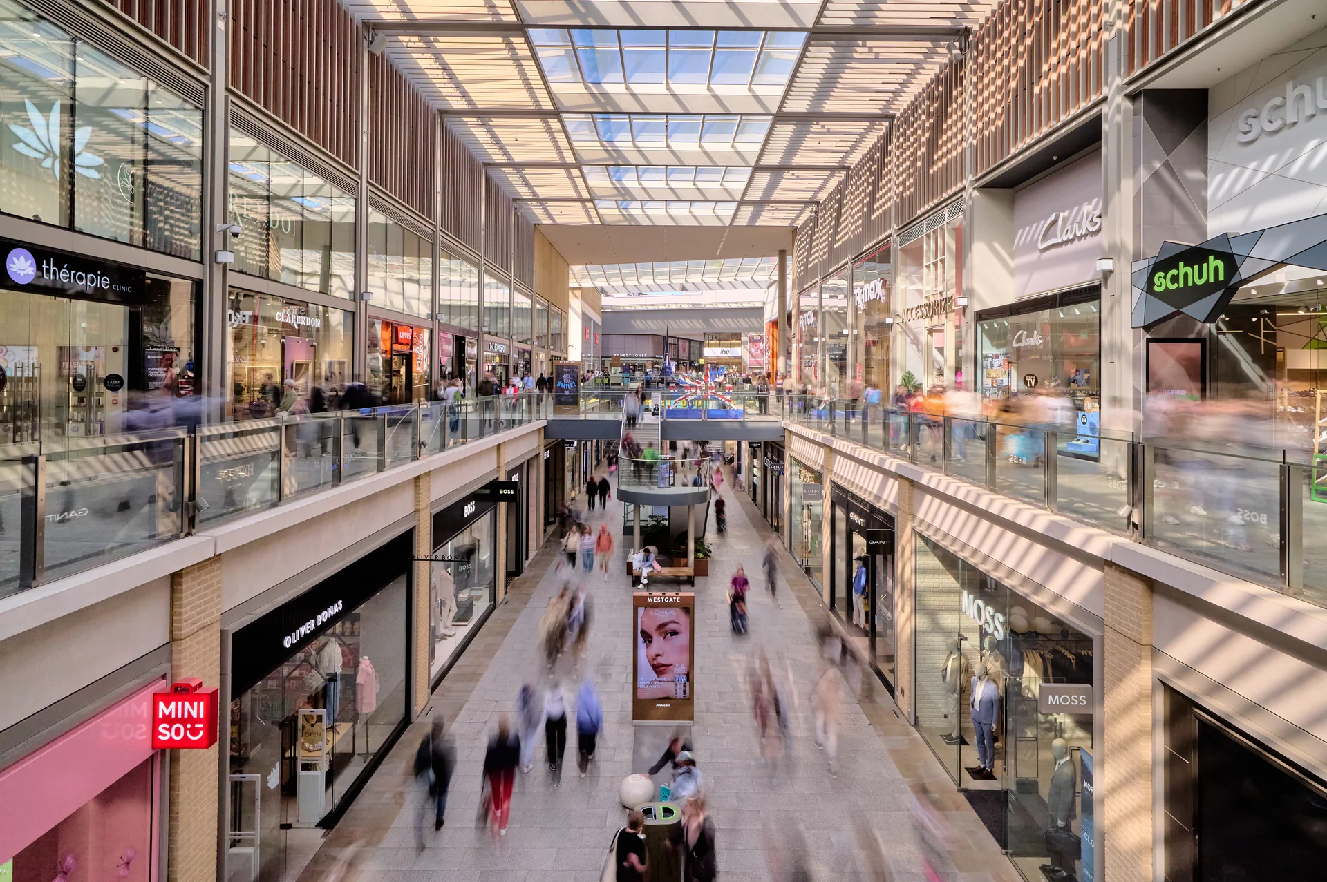 A bustling indoor shopping mall with glass ceilings, multiple levels, and shoppers moving between stores like Mini Sou and Moss.