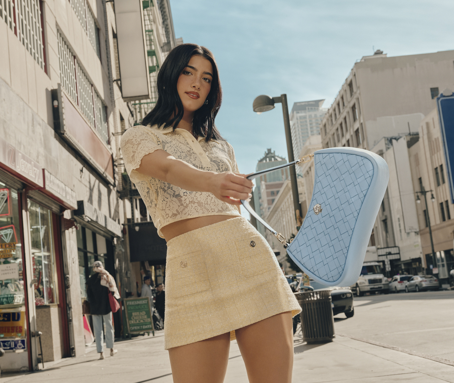 Woman with dark hair and a yellow top and skirt swinging a baby blue Kate Spade New York shoulder bag on a sunny street in a city.
