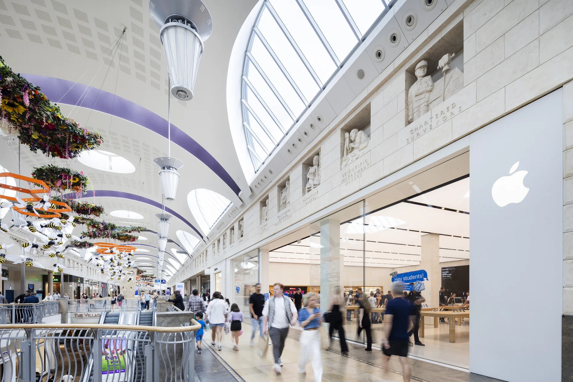 Interior walkway of **Bluewater Shopping Centre** featuring the **Apple Store** entrance, decorative stone carvings above the shops, and overhead floral and bee-themed decorations under a large arched skylight.