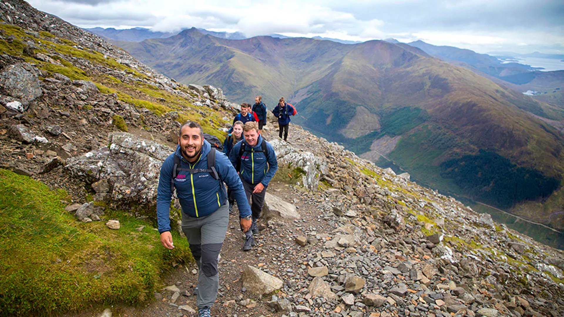 Hikers on a rocky mountain trail with a valley and water in the background.