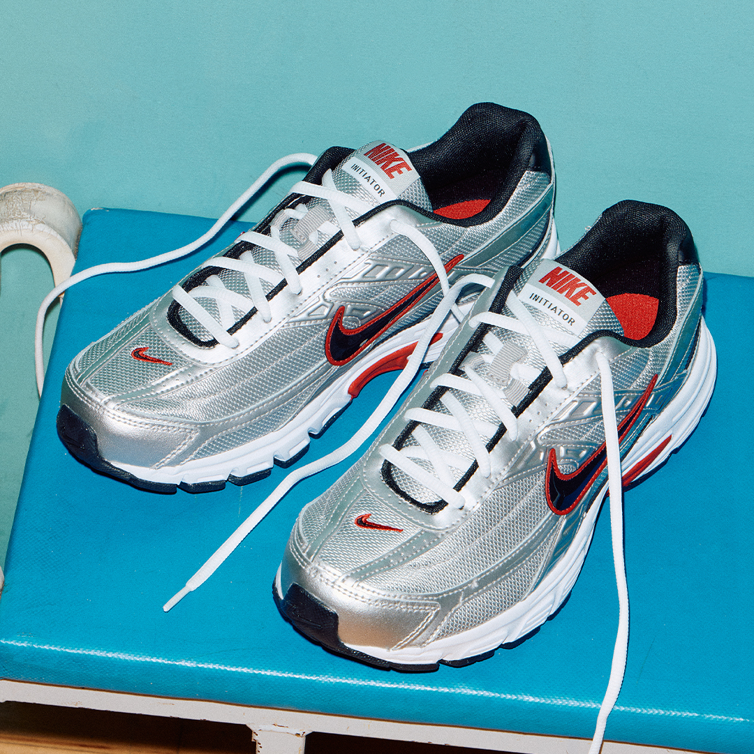 A pair of silver Nikes trainers with laces undone, red on soles and black nike ticks with a red outline, placed on a blue bench 