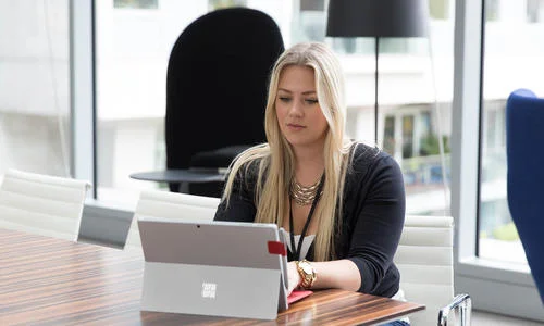 Person with blonde hair using a laptop with a red cover at a wooden table in a modern office.