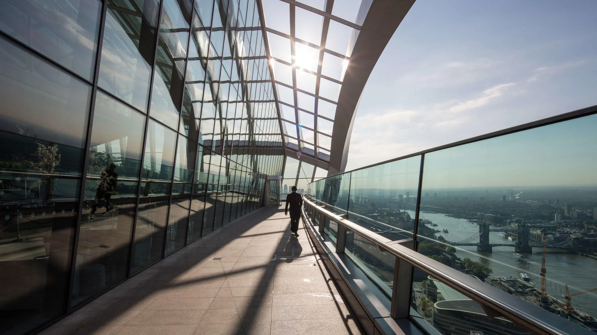 Person walking in a glass-enclosed walkway overlooking a cityscape and river.