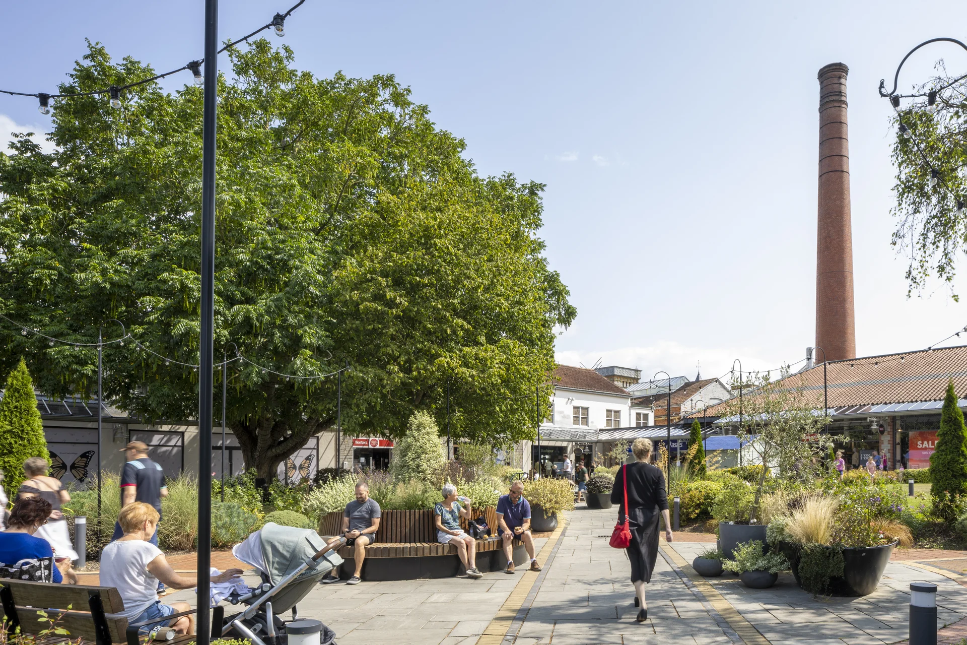 A sunny urban plaza with people relaxing on benches, a woman walking, greenery, a tall chimney, and buildings in the background.