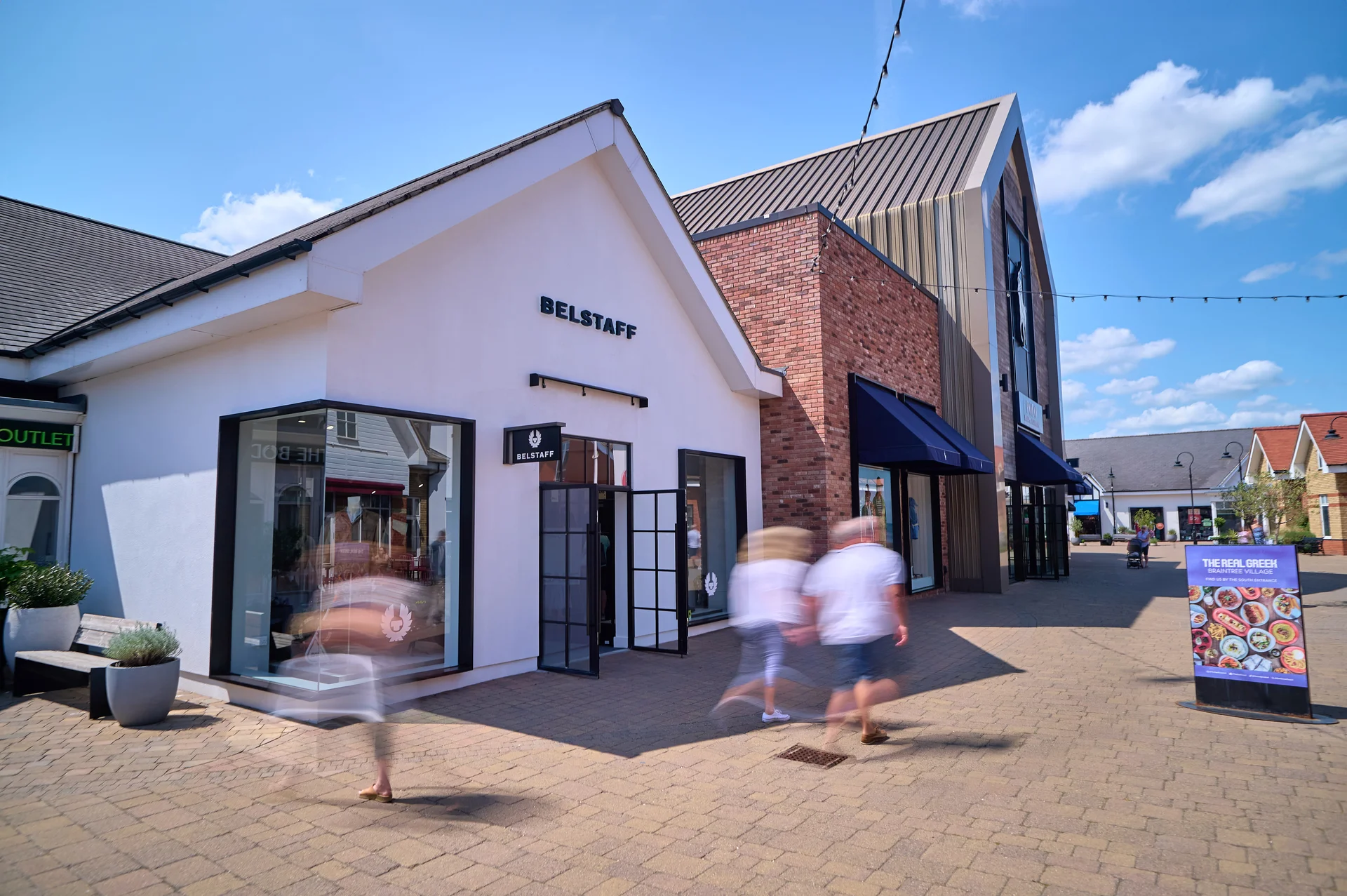 Exterior view of the **Belstaff** store at **Braintree Village** outlet on a sunny day, showcasing its white and brick architecture with motion-blurred shoppers walking on the paved walkway.