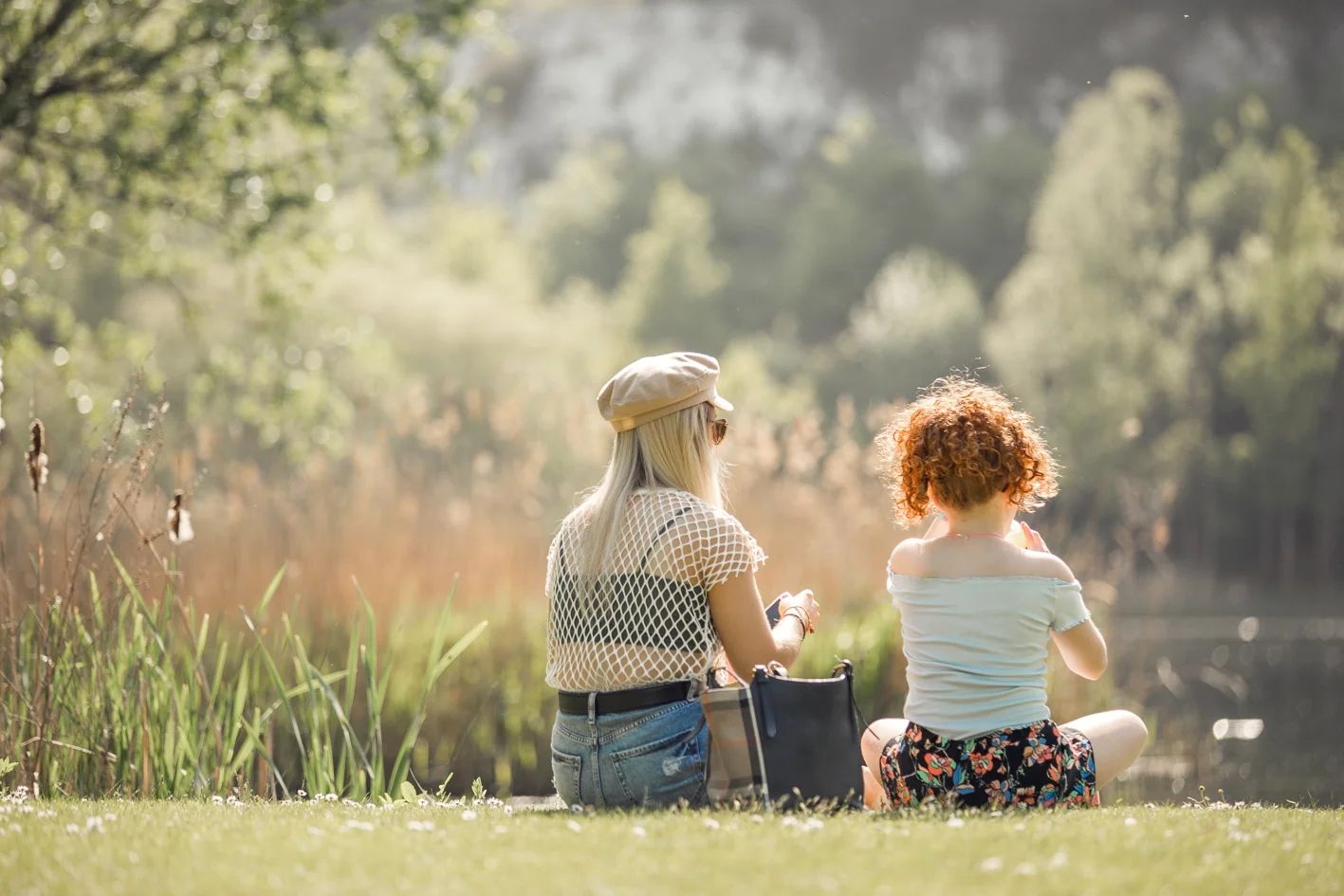 Two people sit on grass by a pond, enjoying a sunny day surrounded by greenery and reeds.